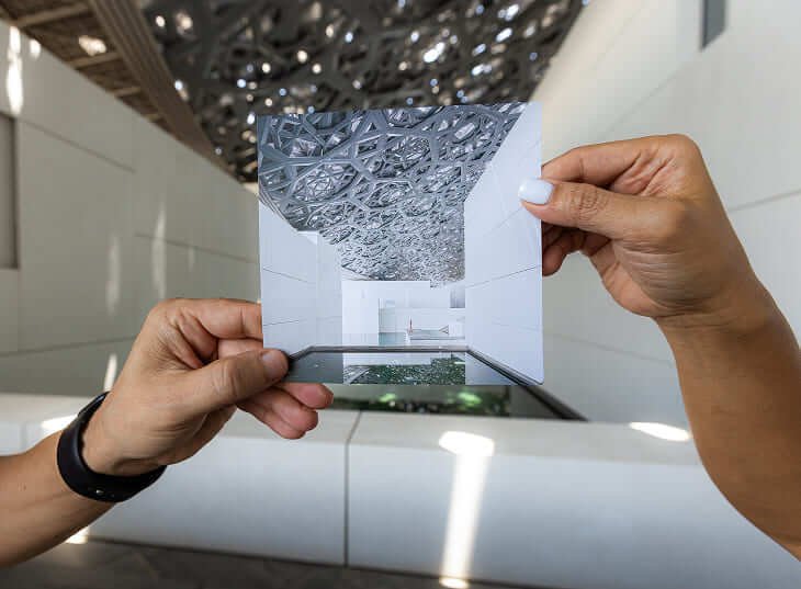 Two hands hold a photo of intricate, geometric ceiling art, aligning it with a real, similar ceiling backdrop. The scene conveys creativity and symmetry.