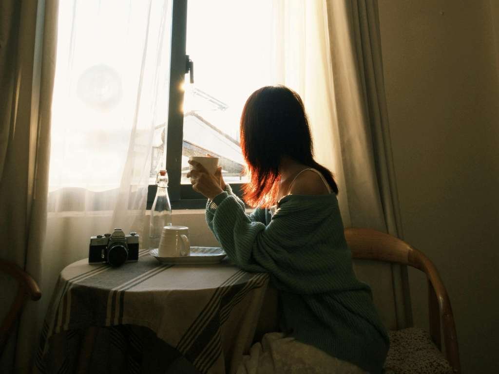 A woman sits by a window holding a cup, looking outside as sunlight falls across the table beside a camera and a bottle.