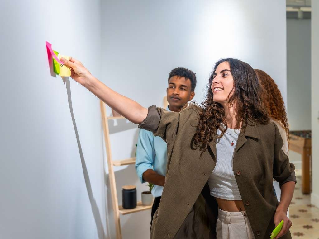 A woman places colorful sticky notes on a wall in a bright office. She looks engaged and happy. Two people watch, showing collaboration and teamwork.