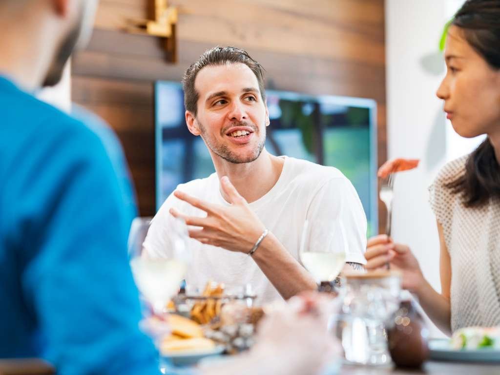 Three people sitting at a table, mid-conversation, talking over food and drinks.